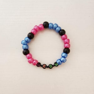 May include: A circular beaded bracelet featuring alternating pink, blue, and black pony beads. The bracelet includes four small black letter beads spelling out the word MEOW in colorful lettering, integrated into the circular design.