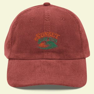 May include: A maroon corduroy baseball cap featuring an embroidered graphic on the front. The design includes the text "SUNSET SURF CLUB" in orange and green, positioned above an orange sun setting behind a stylised green ocean wave.