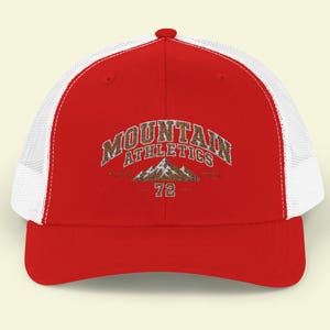 Può includere: Un cappellino trucker rosso e bianco con retro in rete e pannello frontale solido. La parte anteriore mostra il testo MOUNTAIN ATHLETICS in un carattere marrone testurizzato sopra un grafico di una catena montuosa e il numero 72.