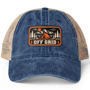 Op de afbeelding: Een blauwe denim baseballpet met een beige mesh achterkant. De voorkant heeft een rechthoekige patch met een oranje en zwarte afbeelding van een tent, bergen en dennenbomen, met de tekst OFF GRID in dikke witte letters onderaan.