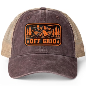 Op de afbeelding: Een bruine baseballpet met een beige mesh achterkant en een rechthoekige patch op de voorzijde. De patch bevat een oranje afbeelding van een tent, bergen, bomen en een zon, met de tekst OFF GRID in dikke, oranje hoofdletters eronder gedrukt.