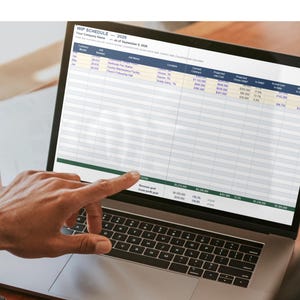 May include: A silver laptop displaying a WIP SCHEDULE 2025 spreadsheet with columns for contract month, job number, job name, location, and financial data. The screen shows rows of project information and a dark header row with blue text.