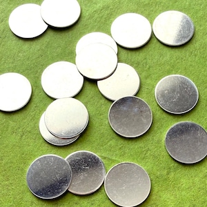 May include: A collection of small, silver-colored, round metal discs scattered on a green surface. The discs appear to be blank and could be used for various craft or jewelry projects. The lighting highlights the metallic sheen of the discs.