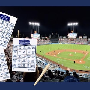 May include: Two white bingo cards with blue text and the Dodgers logo, featuring baseball-themed squares like "Broken Bat" and "Grand Slam." A wooden pencil rests diagonally across the cards, which are set against a night view of a baseball stadium.