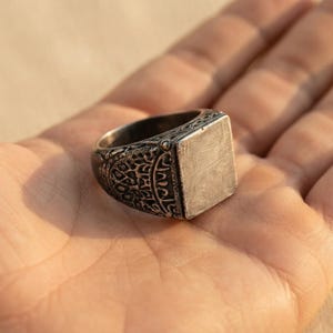 May include: A silver-toned metal ring featuring a flat, rectangular face with a brushed texture. The band displays an intricate, dark, recessed pattern of interconnected shapes and lines that contrast with the polished, smooth surface of the face.