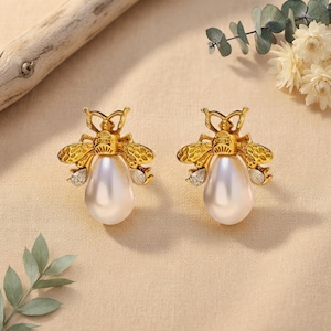 May include: A pair of gold-toned bee-shaped stud earrings featuring textured wings and a central teardrop-shaped white pearl body. Each earring includes small clear accent stones on the lower wing sections, creating a detailed insect design.