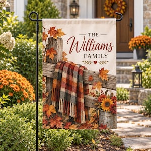 Peut inclure: Un drapeau de jardin rectangulaire représentant une clôture en bois rustique drapée d'une couverture à carreaux rouges, orange et noirs. Le texte indique THE Williams FAMILY avec un petit cœur rouge. Des feuilles d'érable et un seau de tournesols.