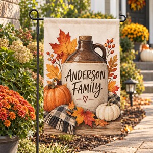 Op de afbeelding: Een rechthoekige tuin vlag met een keramische kruik met de tekst Anderson Family en een hartje. Het ontwerp bevat oranje pompoenen, een zwart-wit geruite deken en kleurrijke herfstachtige esdoornbladeren op een lichte achtergrond.