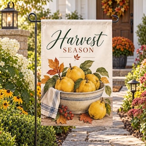 Puede incluir: Una bandera de jardín rectangular con el texto Harvest Season en letra cursiva verde oscuro y marrón. El diseño muestra un cuenco de cerámica lleno de membrillos amarillos, hojas verdes y un paño de rayas azules sobre una superficie de madera.