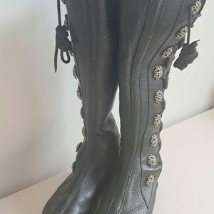 May include: A pair of black knee-high boots featuring a textured leather finish, a rounded toe, and a vertical row of decorative silver-toned metal buttons along the outer side. The boots include black cord lacing details and a thick, lugged sole.