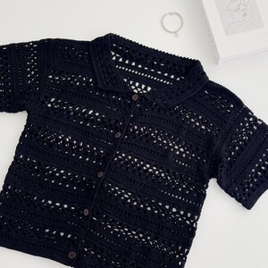 Peut inclure: Une chemise à manches courtes en crochet noir dotée d'un col et d'une fermeture boutonnée sur le devant. Le vêtement est réalisé avec un motif ajouré répétitif de bandes horizontales, créant un aspect texturé et semi-transparent sur l'ensemble.