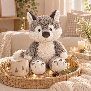 Pode incluir: Um peluche de lobo com pelo texturizado cinzento e creme, orelhas pontiagudas e nariz castanho escuro, sentado ao lado de uma caneca de cerâmica com um padrão repetido de pinheiros. O brinquedo tem patas creme com detalhes de garras castanhas.