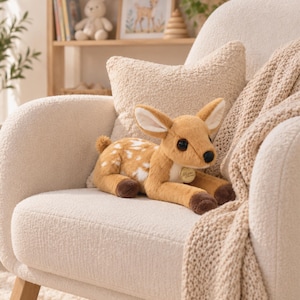 May include: A plush fawn toy with tan fur, white spots on its back, large dark eyes, and dark brown hooves. The stuffed animal features a small round gold-coloured tag attached to its neck area, resting in a seated position on a textured fabric.