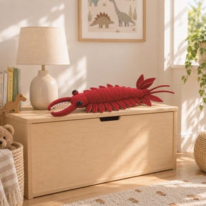 Könnte beinhalten: Ein tiefrotes, segmentiertes Plüschtier, das einem Anomalocaris ähnelt, mit großen schwarzen Augen, mehreren seitlichen Flossen und einem langen, spitzen Schwanz. Das Wesen hat zwei markante, gebogene Vorderanhänge mit kleinen weißen Zähnen.