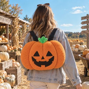 Könnte beinhalten: Ein orangefarbener Plüsch-Kürbis-Rucksack mit einem schwarzen Jack-o'-Lantern-Gesicht aus Filz, bestehend aus dreieckigen Augen und einem gezackten Mund, gekrönt von einem kleinen grünen Stiel. Der Rucksack hat schwarze, verstellbare Träger.
