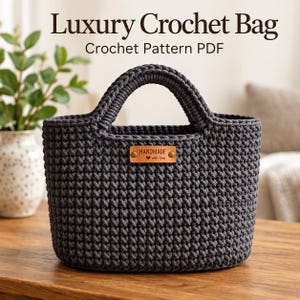 May include: A dark gray crochet bag with two integrated handles and a rectangular tan leather tag centered on the front. The tag features the text "HANDMADE" with a small heart icon. The text "Luxury Crochet Bag Crochet Pattern PDF" is visible.