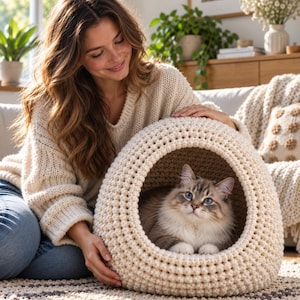 May include: A cream-colored, round, chunky knit cat bed with a wide circular opening. A fluffy, long-haired cat with grey and white fur and blue eyes is resting inside the textured, soft-looking, spherical pet cave made of thick yarn.