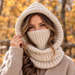 Può includere: Un cappuccio con scaldacollo all'uncinetto color beige con un motivo a maglia spesso e testurizzato. L'accessorio presenta un cappuccio profondo che incornicia il viso e uno scaldacollo alto a coste che copre la parte inferiore del volto.