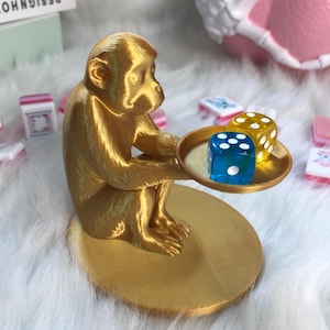 May include: A gold-coloured monkey figurine sitting on a circular base and holding a small round tray. On the tray are two translucent dice, one blue and one yellow, each featuring white pips. The monkey is depicted in a seated position.
