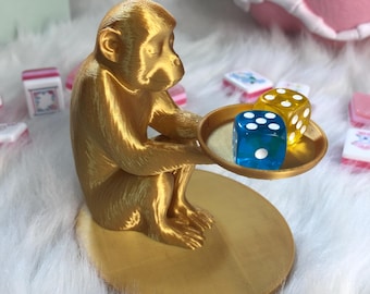 Peut inclure: Une figurine de singe couleur or assise sur une base circulaire et tenant un petit plateau rond. Sur le plateau se trouvent deux dés translucides, un bleu et un jaune, chacun avec des points blancs. Le singe est représenté en position assise.