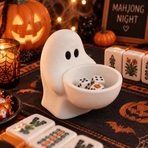 May include: A white ghost-shaped ceramic bowl with two black oval eyes, holding three six-sided dice inside its rounded basin. The dice feature standard black pips, with one die displaying a small orange pumpkin graphic on one of its faces.