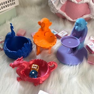 Könnte beinhalten: Eine Sammlung von vier 3D-gedruckten dekorativen Schalen und Ablagen, bestehend aus einem blauen Pfau, einem orangefarbenen Affen mit blattförmiger Schale, einem violetten Affen mit rundem Tablett und einer roten Drachenschale mit zwei Würfeln.