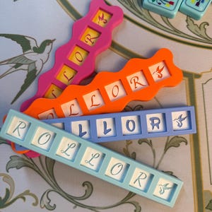 May include: Four rectangular game tiles with wavy edges in pink, orange, light blue, and white. Each tile features square cutouts containing letters that spell ROLLOR, with some tiles displaying a small hummingbird graphic at the end of the sequence.