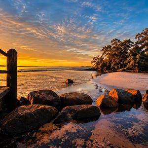 May include: A wooden pier extends into a calm body of water at sunset, featuring dark brown wooden posts and planks. Large, smooth, dark grey rocks are scattered in the shallow water near a sandy beach lined with dense green trees under a golden sky.