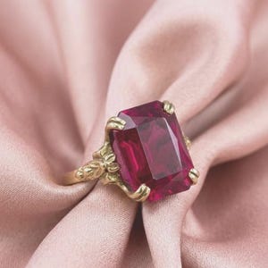 May include: A gold ring featuring a large, rectangular, deep red gemstone set in a claw-style mounting. The metal band displays intricate, textured detailing near the setting, which securely holds the faceted stone in a prominent, elevated position.