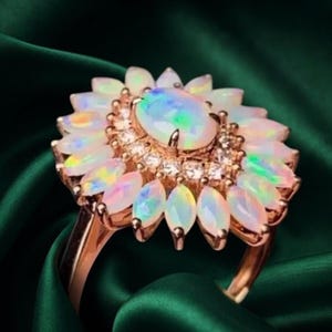 May include: A rose gold ring featuring a central oval opal surrounded by a halo of small clear stones and an outer layer of marquise-cut opals arranged in a floral pattern. The opals display iridescent flashes of green, blue, and pink colors.