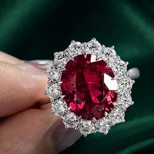 Op de afbeelding: Een ring met een grote, ovaal geslepen dieprode edelsteen in het midden, omringd door een halo van talloze kleine, ronde, helderwitte stenen. De zetting is van metaal en het ontwerp creëert een bloemachtig uiterlijk met de stenen.