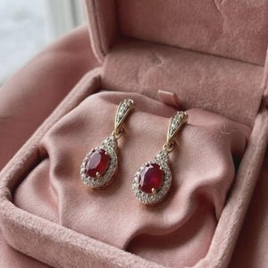 May include: A pair of gold drop earrings featuring oval-cut deep red rubies surrounded by a halo of small white diamonds. The earrings have a tapered top section also set with small white diamonds, creating a teardrop-shaped design.