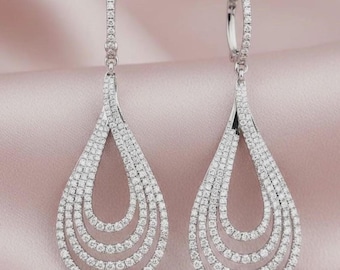 May include: A pair of silver-toned drop earrings featuring a teardrop shape composed of three concentric open loops. Each loop is encrusted with small, clear, sparkling stones along the entire surface, including the vertical post attachment.