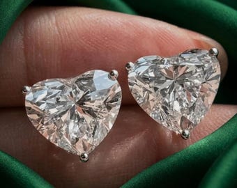 May include: A pair of heart-shaped stud earrings featuring clear, faceted stones set in silver-toned metal prongs. The stones exhibit a brilliant, reflective quality with multiple sharp facets that catch the light, creating a sparkling appearance.