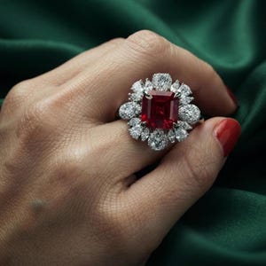 Könnte beinhalten: Ein Ring mit einem großen, rechteckigen, tiefroten Edelstein in der Mitte, umgeben von einem Kranz aus mehreren kleineren, klaren, birnenförmigen und rund geschliffenen weißen Steinen in einer Metallfassung, die blumenartig angeordnet sind.