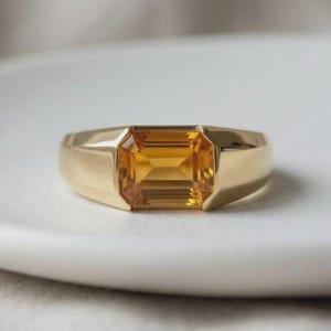 Peut inclure: Une bague en or ornée d'une pierre précieuse citrine jaune de taille émeraude rectangulaire au centre. L'anneau est lisse et poli, avec une construction en métal solide et large qui s'affine légèrement vers l'arrière de la monture.