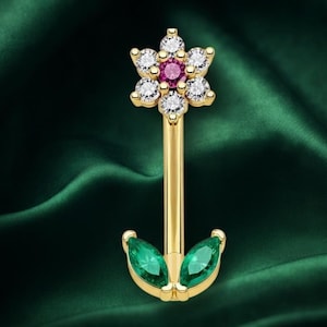May include: A gold curved barbell body jewelry piece featuring a flower-shaped top with five clear round stones surrounding a central pink stone. The bottom end displays two marquise-cut green stones arranged in a leaf-like design on a gold base.