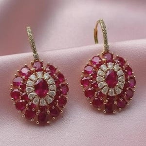 May include: A pair of gold drop earrings featuring a central oval-cut red gemstone surrounded by a halo of small white stones, with an outer layer of larger round-cut red gemstones. The gold ear wires are accented with small white stones.