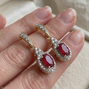 May include: A pair of gold drop earrings featuring oval-cut red rubies surrounded by halos of small white diamonds. The earrings have hinged clasps also accented with tiny diamonds, creating a cohesive and elegant jewelry design.