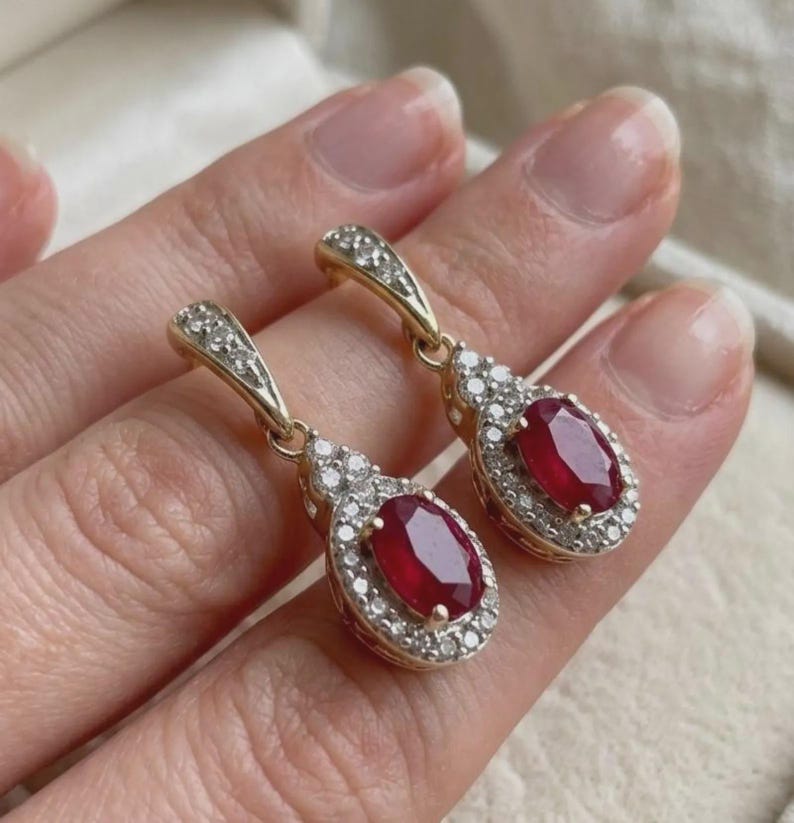 May include: A pair of gold drop earrings featuring oval-cut red rubies surrounded by halos of small white diamonds. The earrings have hinged clasps also accented with tiny diamonds, creating a cohesive and elegant jewelry design.
