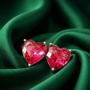 May include: A pair of stud earrings featuring faceted, heart-shaped red gemstones set in silver-toned metal. The stones have a brilliant, reflective surface with multiple geometric facets that catch the light, creating a deep crimson appearance.
