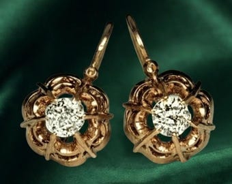 May include: A pair of gold drop earrings featuring a circular, flower-like design with a central round, clear faceted stone. Each earring is secured with a curved wire hook, showcasing a polished metallic finish and intricate petal-like detailing.
