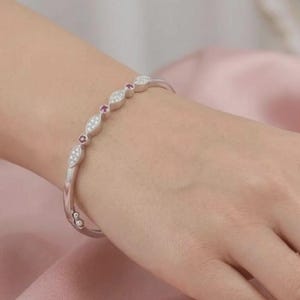 May include: A silver-toned bangle bracelet featuring a series of marquise-shaped links set with small clear stones, alternating with small round red stones. The bracelet has a hinged clasp mechanism and a polished metallic finish throughout.