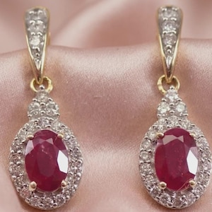May include: A pair of drop earrings featuring oval-cut red rubies surrounded by halos of small white diamonds. The earrings are set in gold metal with additional diamonds embedded along the upper hook and the connecting bail.