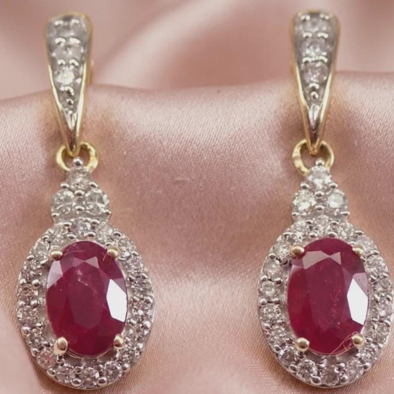 May include: A pair of drop earrings featuring oval-cut red rubies surrounded by halos of small white diamonds. The earrings are set in gold metal with additional diamonds embedded along the upper hook and the connecting bail.