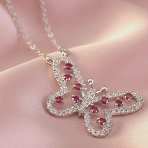 May include: A silver-toned necklace featuring a butterfly-shaped pendant. The pendant is outlined with small clear stones and accented with several round, deep pink-red stones set within the wings. The chain is a delicate, thin silver-toned link.