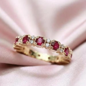 May include: A gold band ring featuring a repeating pattern of round red rubies and small clear white diamonds. The metal setting has a textured, milgrain-style edge around each gemstone, creating a continuous, detailed design along the entire band.