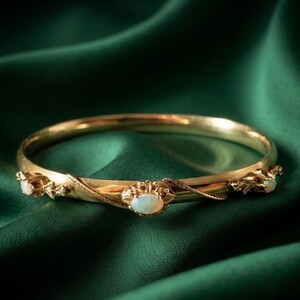 Può includere: Un bracciale rigido in oro caratterizzato da una fascia liscia e lucida, decorata con tre opali ovali e iridescenti. Gli opali sono incastonati in montature metalliche decorative a forma di foglia che avvolgono la circonferenza del gioiello.