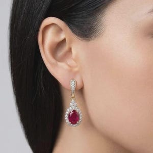 May include: A gold drop earring featuring a central oval-cut red ruby gemstone surrounded by a halo of small white diamonds. The earring includes a decorative bail also set with small white diamonds, suspended from the earlobe.