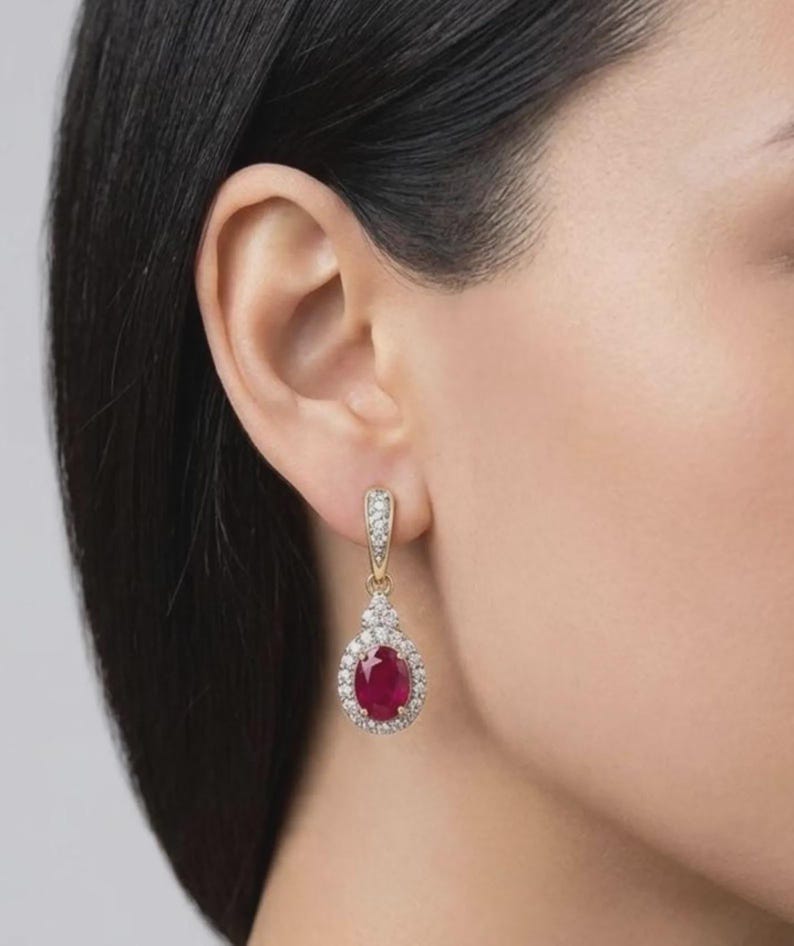 May include: A gold drop earring featuring a central oval-cut red ruby gemstone surrounded by a halo of small white diamonds. The earring includes a decorative bail also set with small white diamonds, suspended from the earlobe.